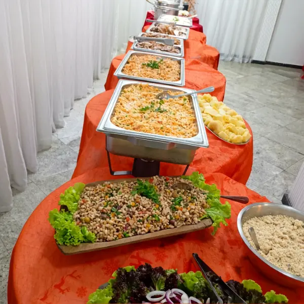 : Buffet servido em mesa com toalha laranja, contendo pratos quentes em travessas de aço e saladas